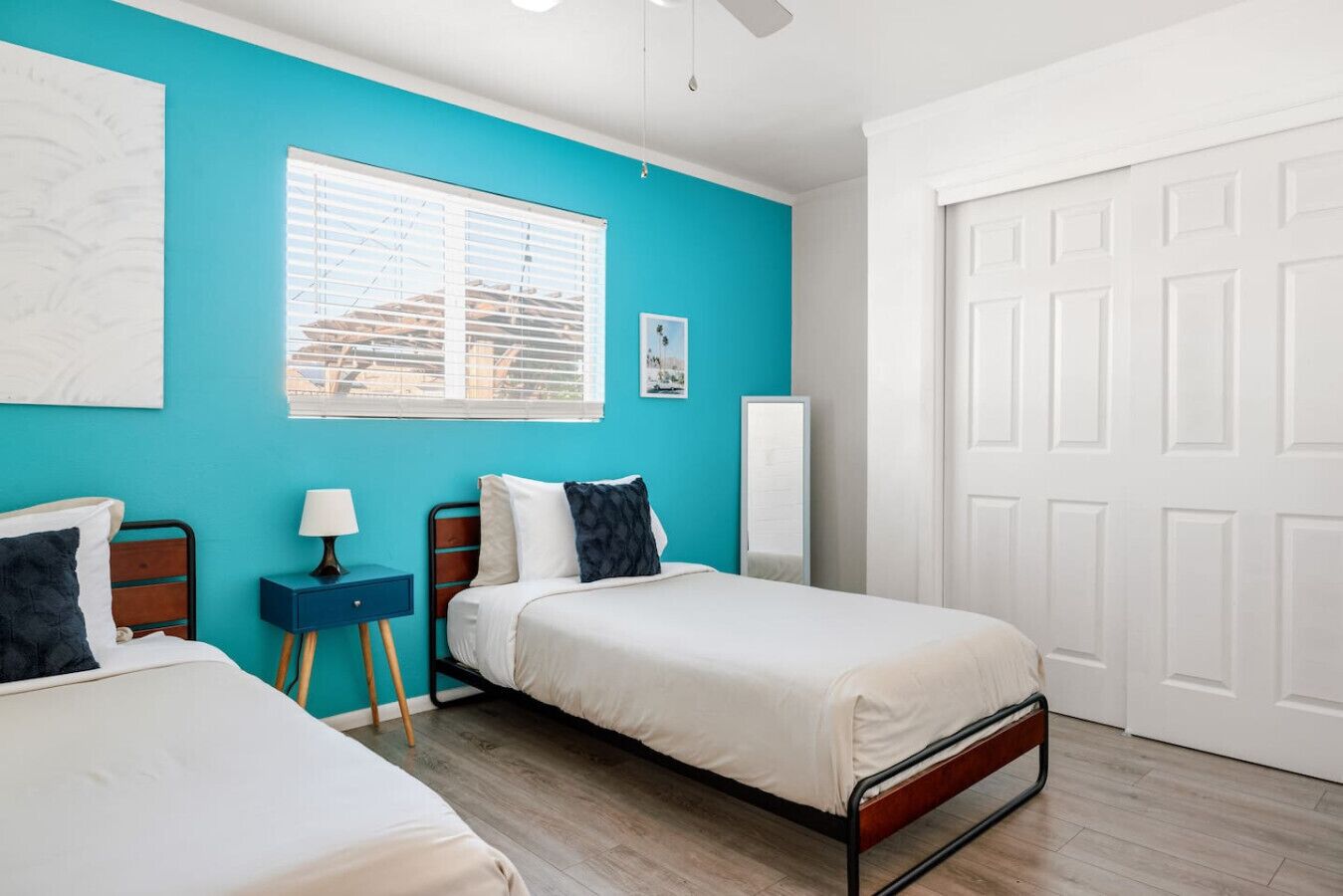 A bedroom with two beds, a window with blinds, a white door, and a small bedside table with a lamp.