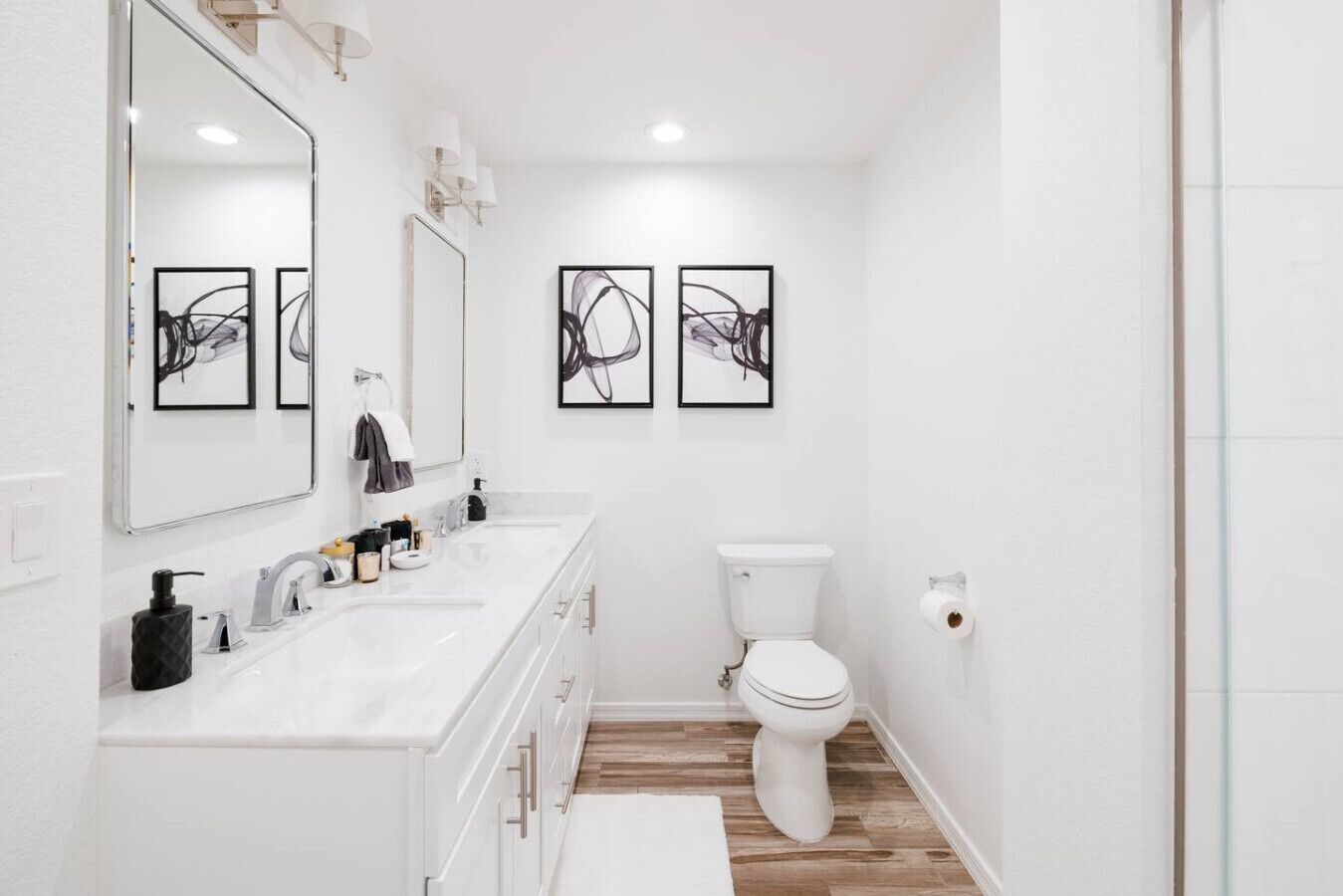 A modern bathroom with a white vanity, two sinks, a toilet, and abstract artwork on the walls.