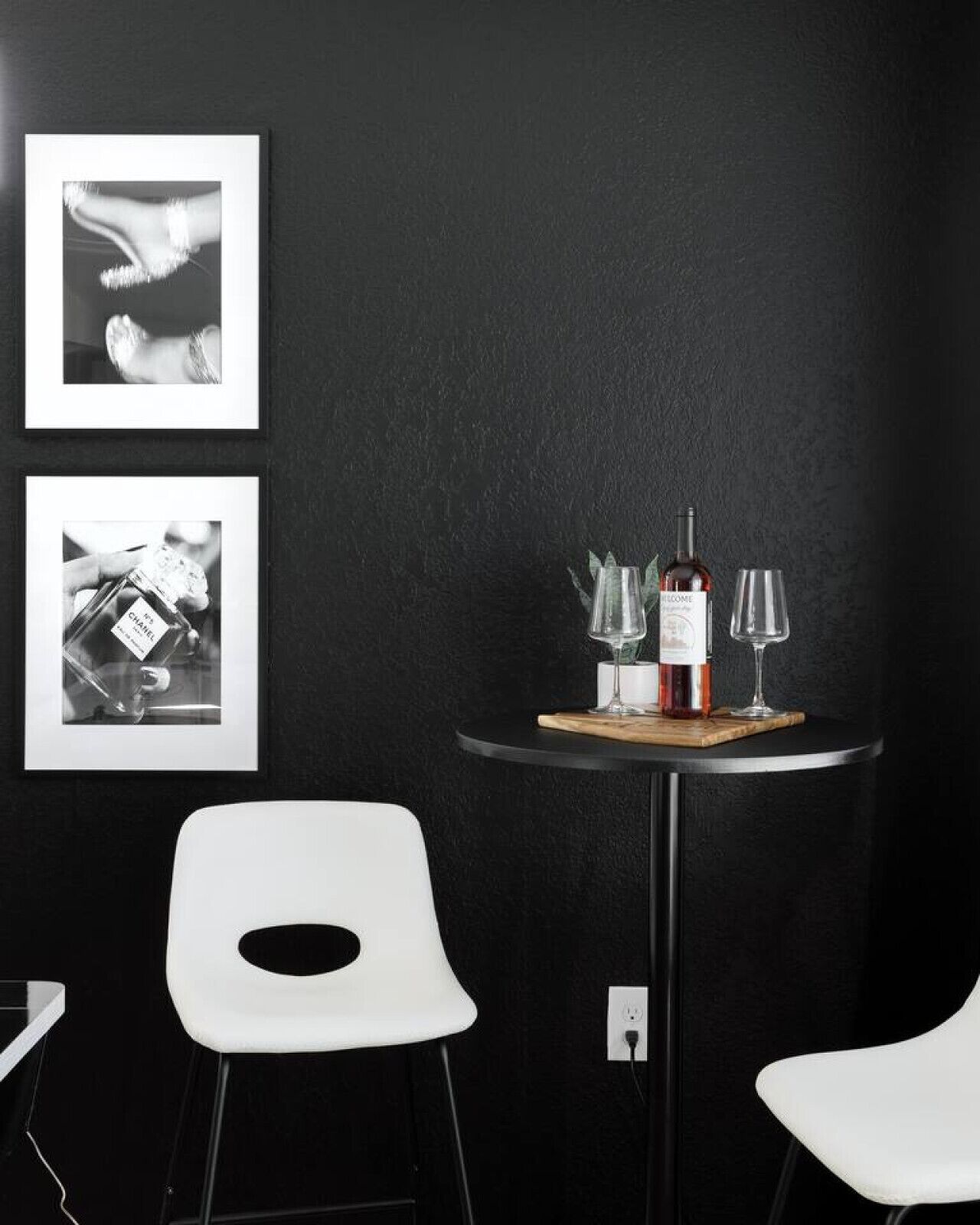 A modern room with a black wall, two white chairs, a small round table with a bottle of wine, two glasses, and two framed pictures.