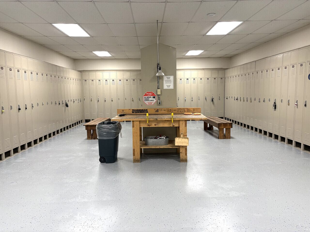 A locker room with a wooden workbench, trash bin, and a sign indicating surveillance.
