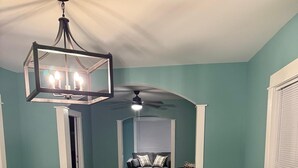 Un comedor con mesa y sillas de madera, un ventilador de techo y vista a una sala de estar con sofá y ventana.
