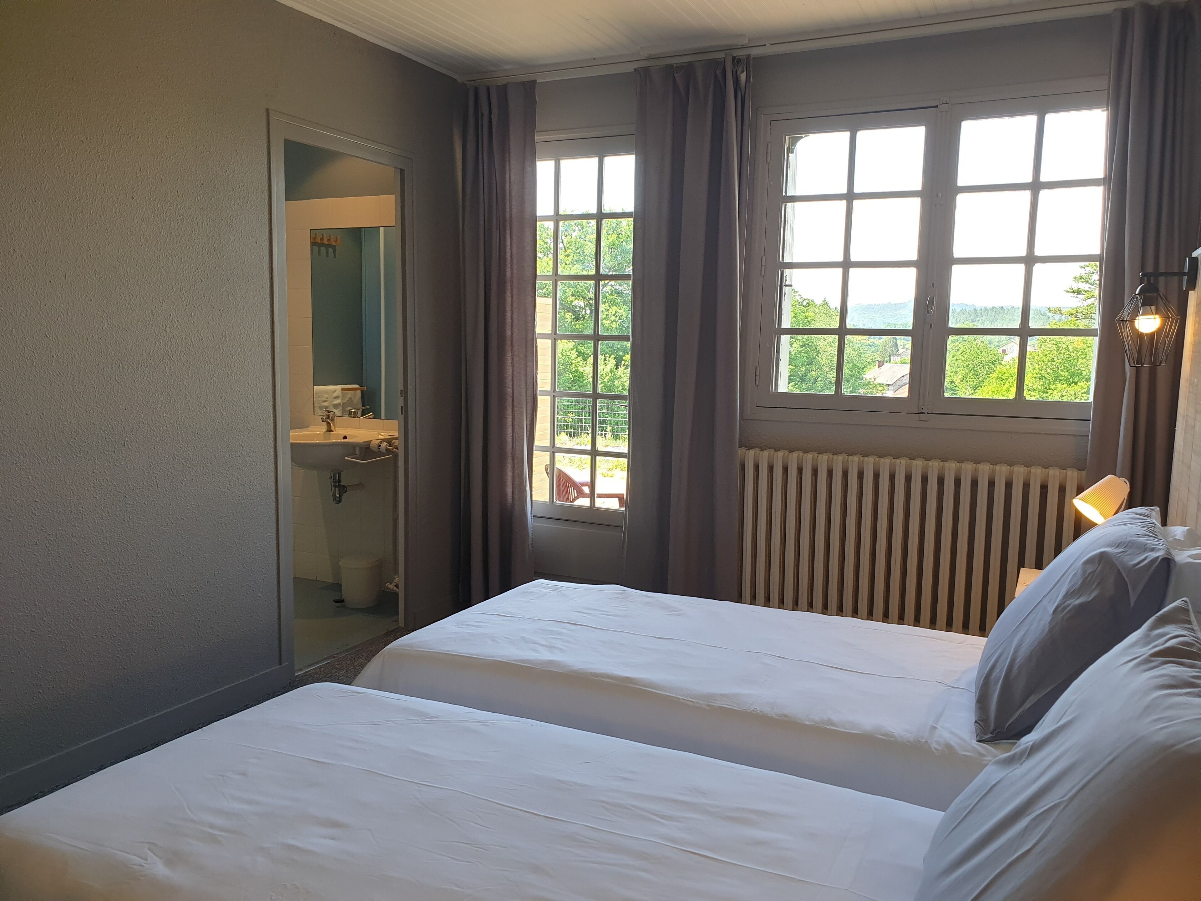 Quarto com duas camas, janela com cortinas, radiador e banheiro visível por uma porta aberta.