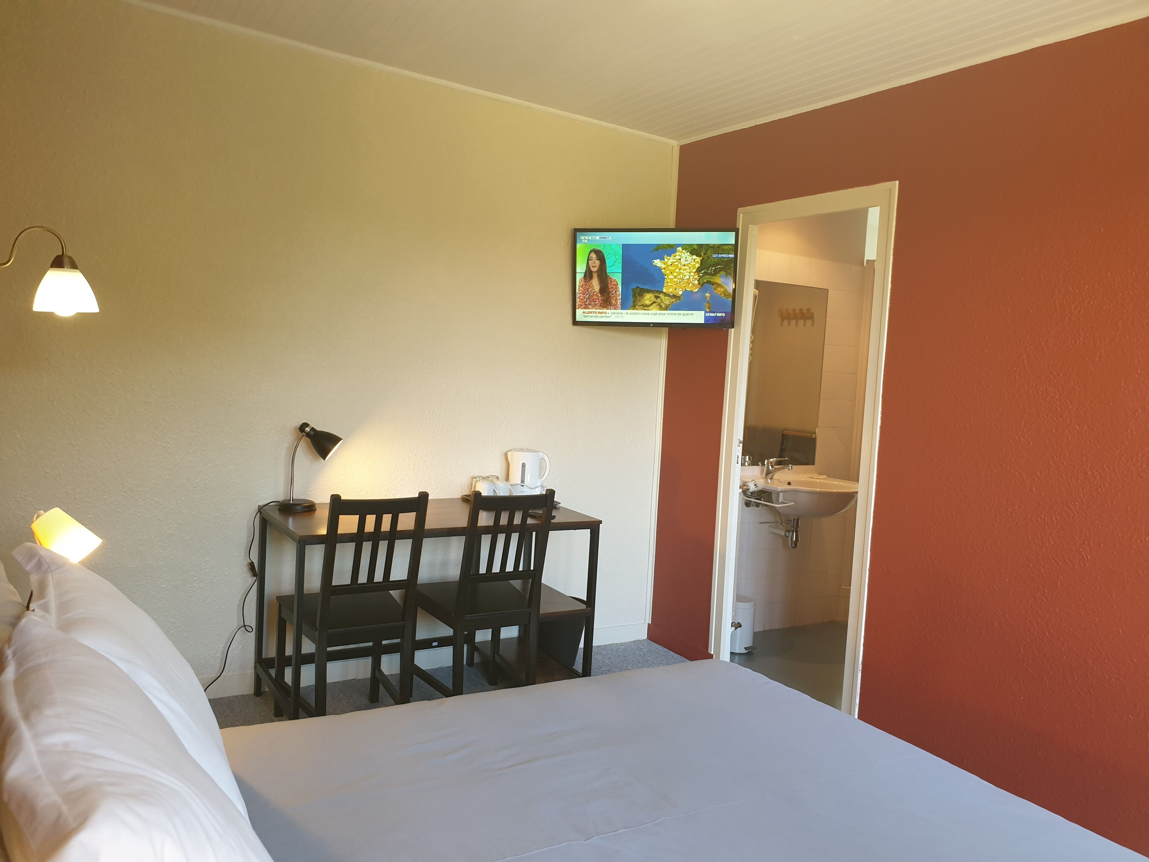A hotel room with a bed, a desk with a chair, a television, and a bathroom visible through an open door.
