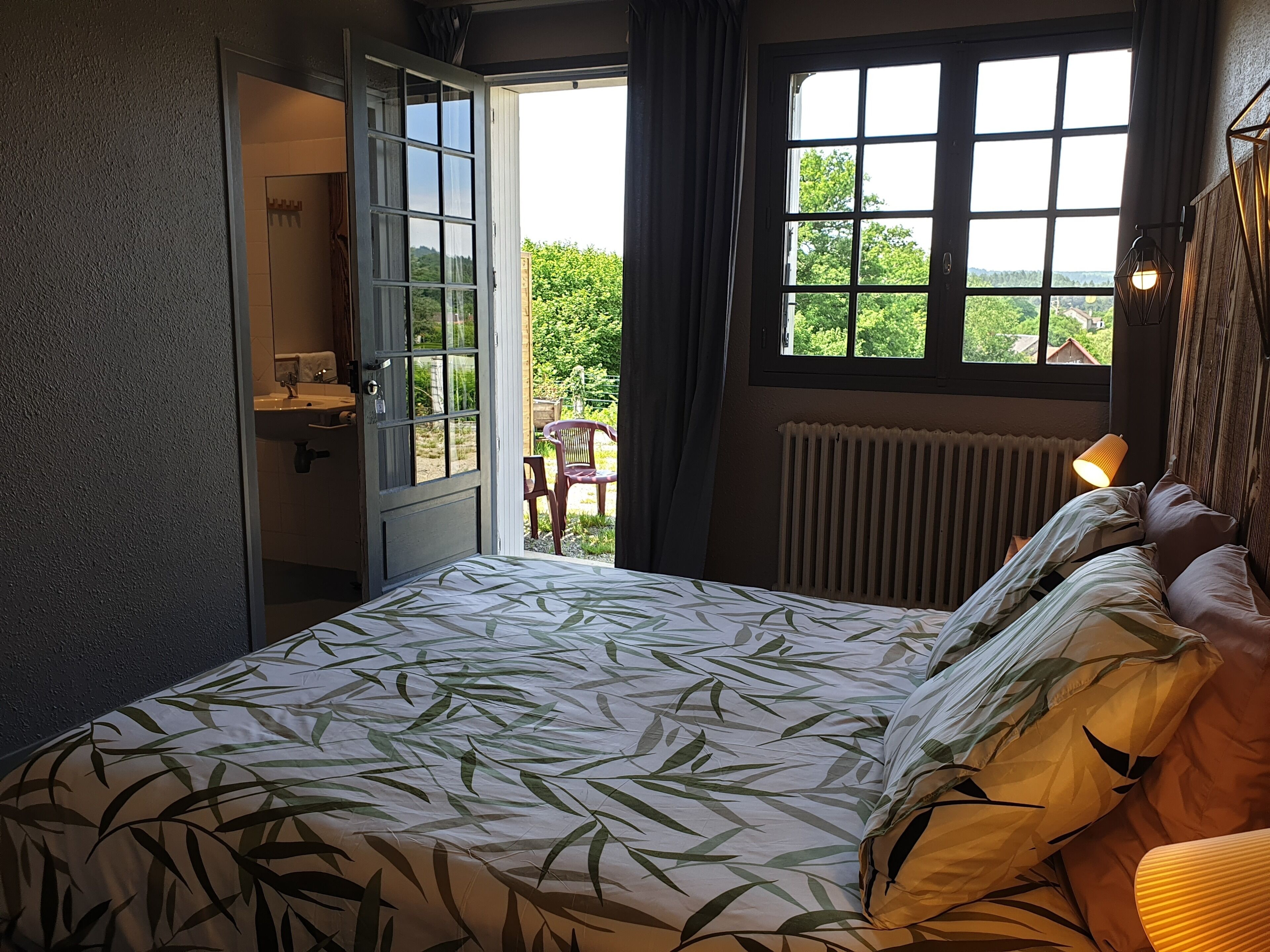 Une chambre à coucher avec un lit, une fenêtre donnant sur un espace verdoyant, un radiateur et une porte menant à la salle de bain.