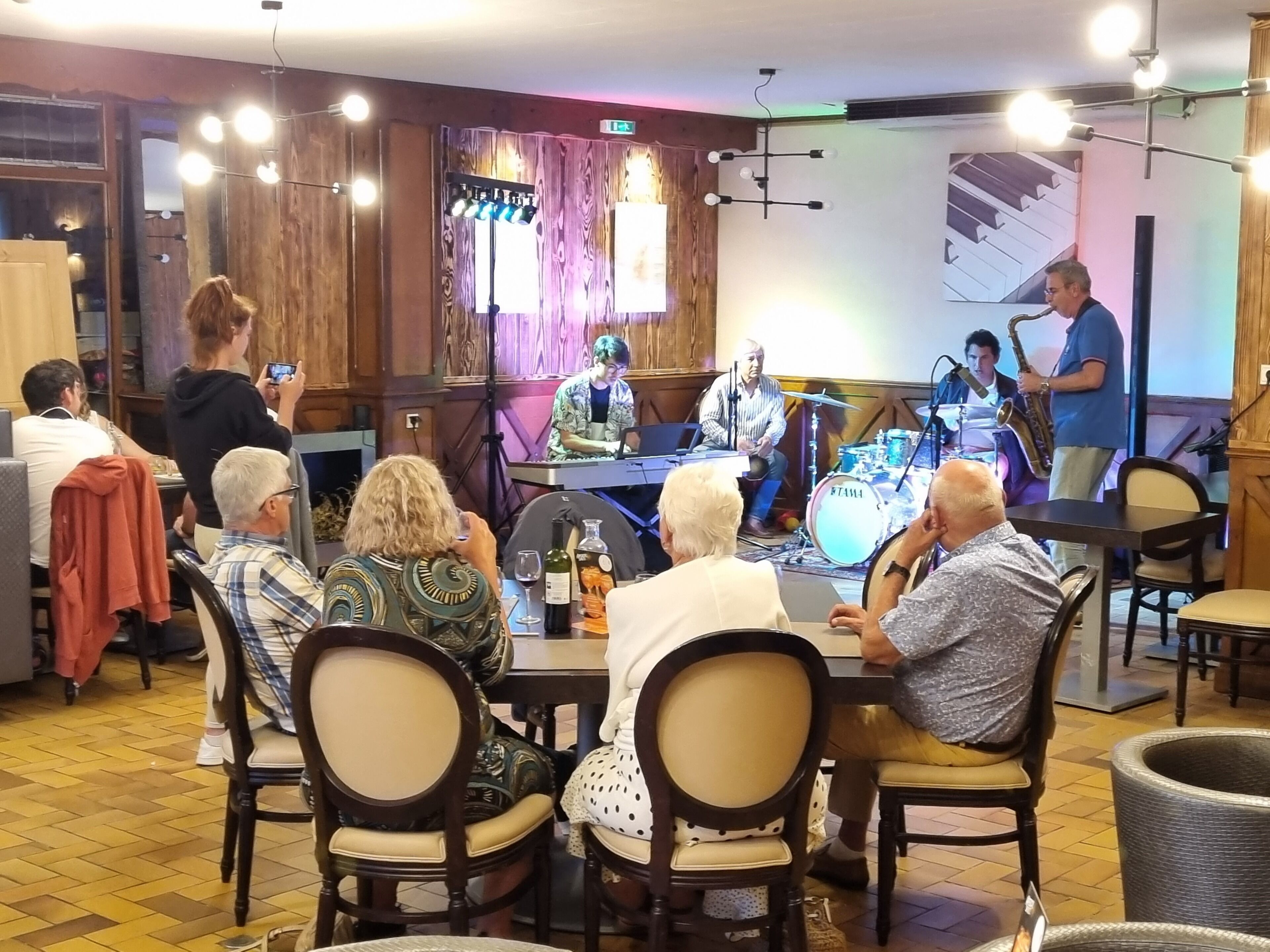 Un groupe de personnes est assis à des tables dans une pièce aux murs en bois, avec une scène pour les concerts.
