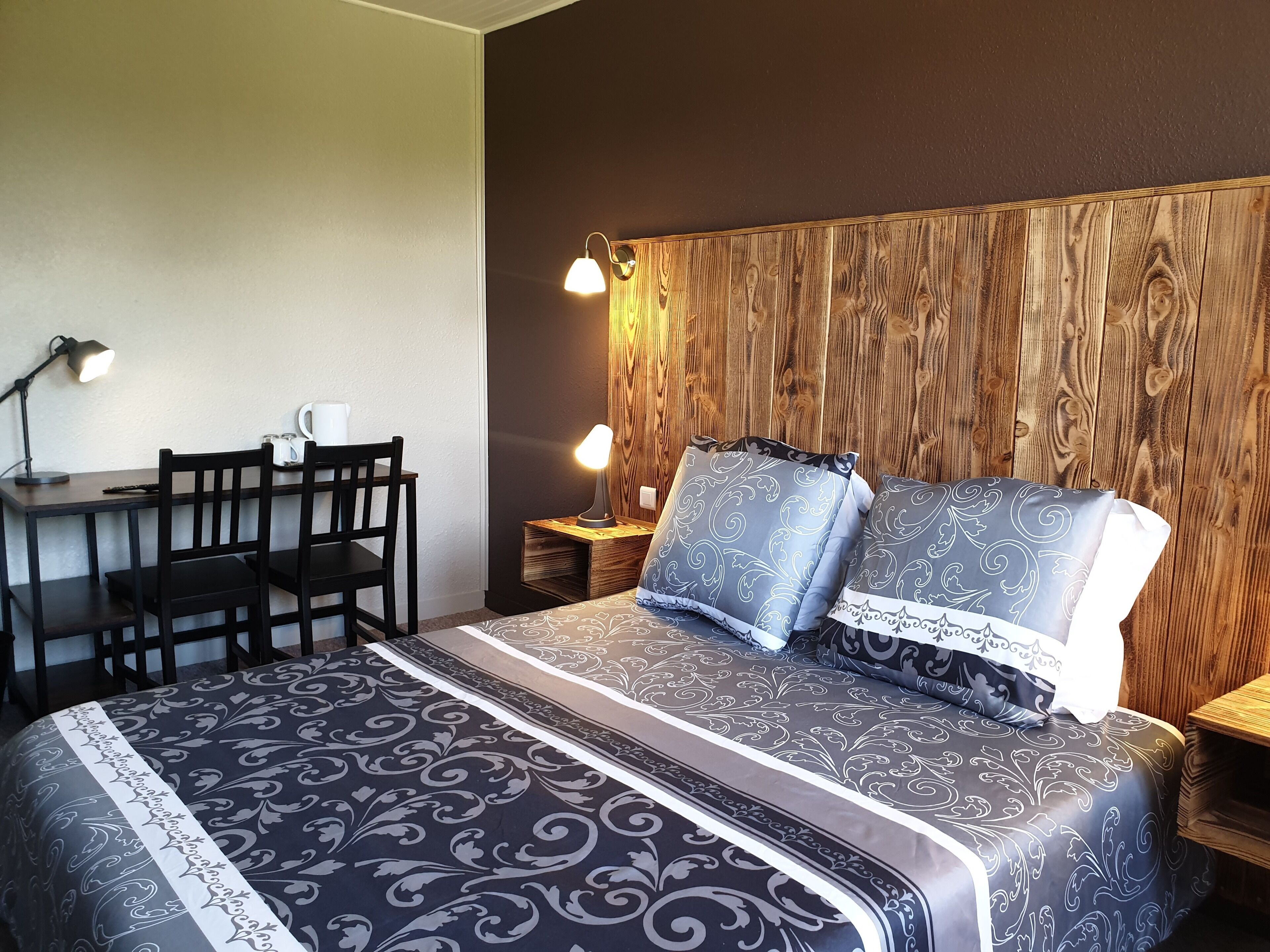 A neatly arranged bedroom with a wooden headboard, a patterned bedspread, and a desk with a chair and lamp.