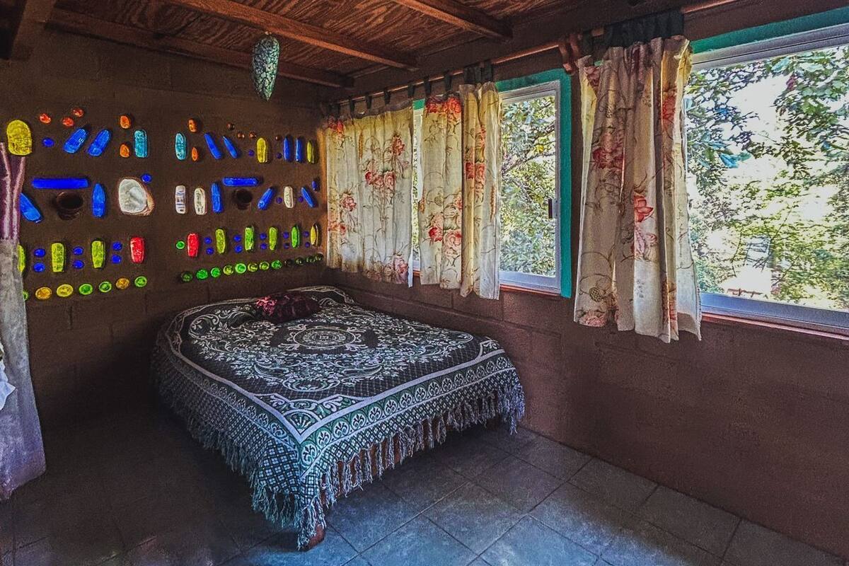 Una stanza con un letto, una parete di bottiglie di vetro colorate e una finestra con tende floreali.