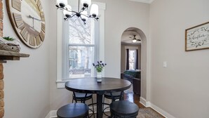 A dining area with a round table, four chairs, a chandelier, a large wall clock, and a window with blinds. - Downtown Private Studio w/ Kitchenette-near UD (Dayton)