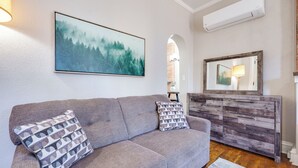 A living room with a gray sofa, two patterned pillows, a painting of a forest, a wooden dresser, and an air conditioning unit. - Downtown Private Studio w/ Kitchenette-near UD (Dayton)
