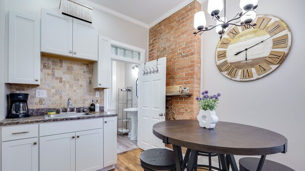 A kitchen with white cabinets, a brick accent wall, a round wooden table with four stools, and a large wall clock. - Downtown Private Studio w/ Kitchenette-near UD (Dayton)