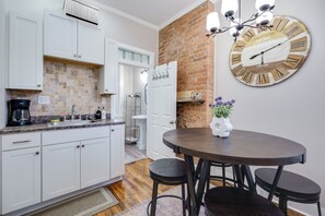 Eine Küche mit weißen Schränken, einer Wand aus Ziegelsteinen, einem runden Holztisch mit vier Barhocker und einer großen Wanduhr.