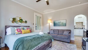 A bedroom with a bed, a gray sofa, a ceiling fan, a painting, and a kitchen visible through an archway. - Downtown Private Studio w/ Kitchenette-near UD (Dayton)
