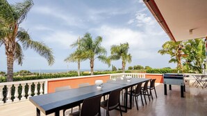 An outdoor dining area with a long table and chairs, surrounded by palm trees and a view of the sea. - Massaroom (Conversano)