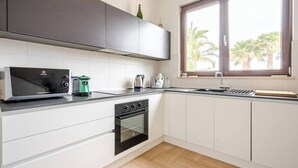 A modern kitchen with white cabinets, a built-in oven, a microwave, and a window with a view of palm trees. - Massaroom (Conversano)
