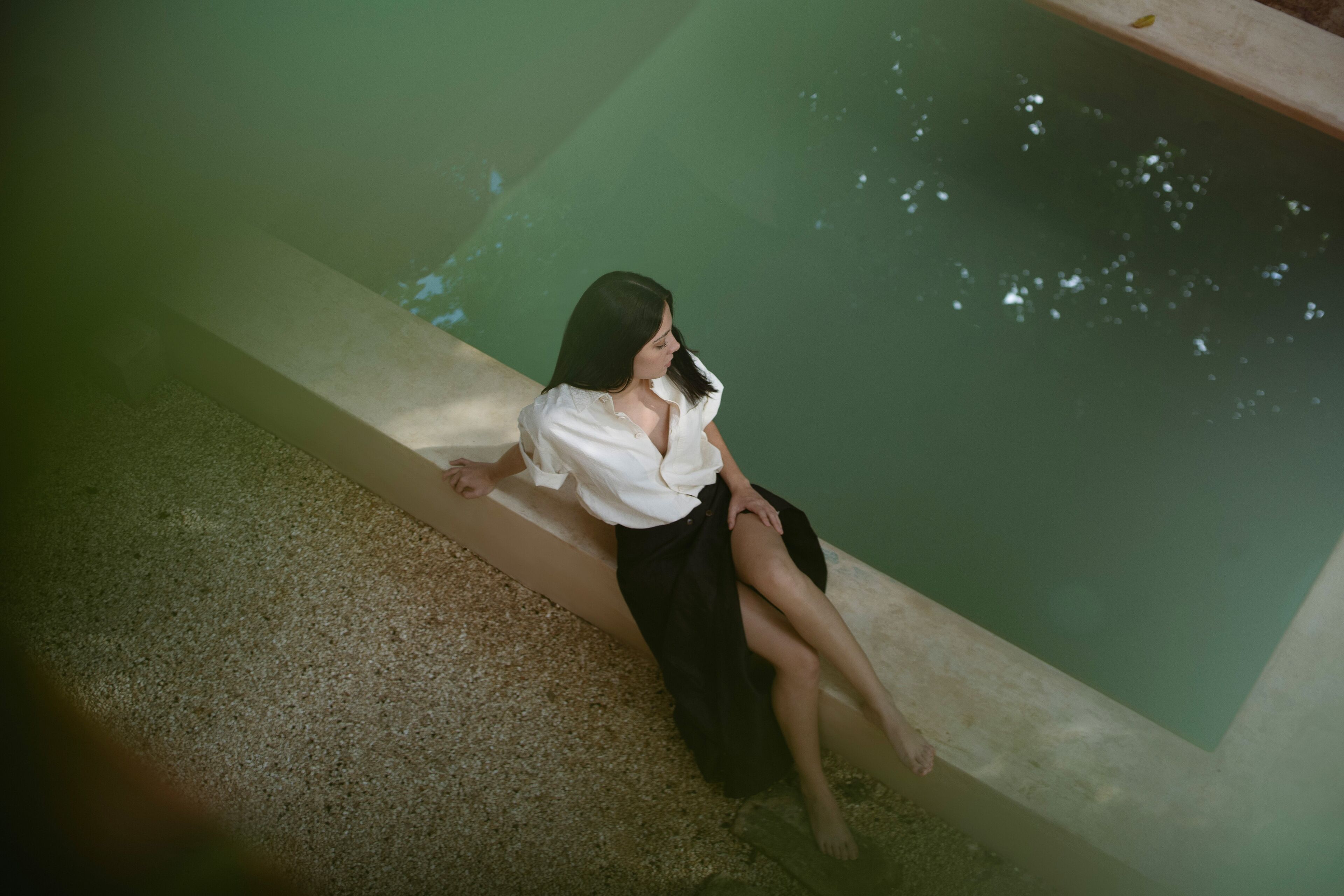 Une personne est assise au bord d’une piscine dont le bord est en béton et le fond recouvert de petits galets.