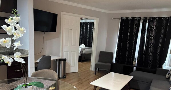A modern hotel room with a flat-screen TV, a dining area with a table and chairs, a sofa, and a large window with patterned curtains.