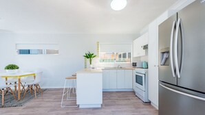 Eine moderne Küche mit weißen Schränken, einem Edelstahlkühlschrank, einem Holztisch mit weißen Stühlen und einer kleinen Kochinsel mit Sitzgelegenheiten.