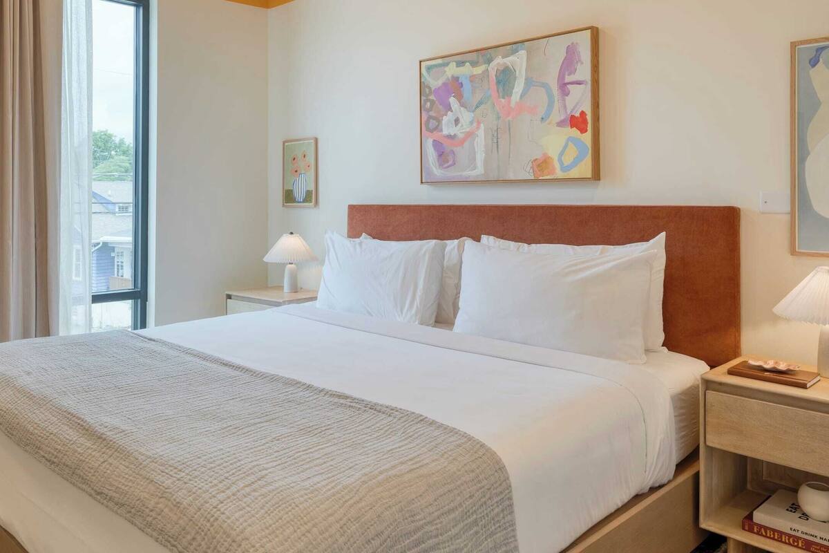 Une chambre d’hôtel avec un grand lit, une table de chevet avec une lampe, un tableau au mur et une fenêtre avec des rideaux.