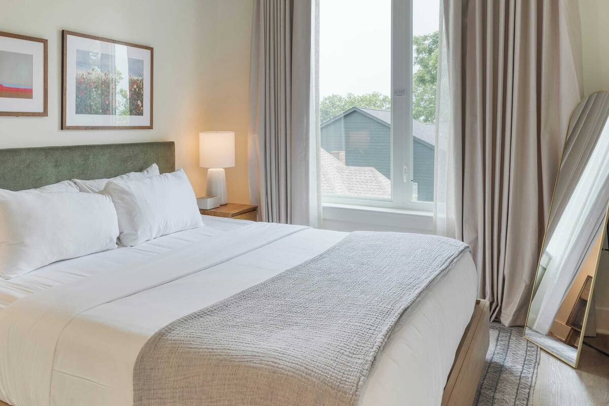 Une chambre d’hôtel avec un grand lit, une table de chevet avec une lampe, deux tableaux encadrés au mur et une fenêtre avec des rideaux.