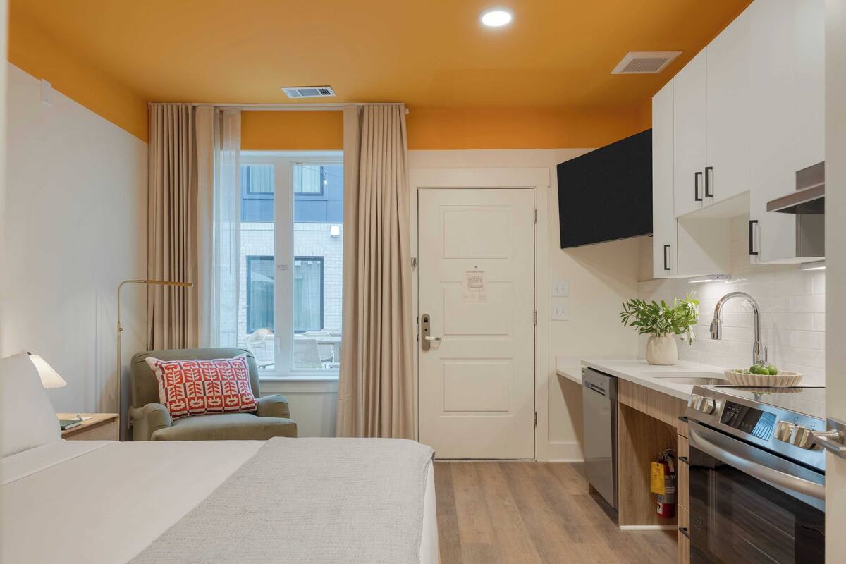 Une chambre d’hôtel de taille réduite, comprenant un lit, un bureau, une chaise, une kitchenette et une vue sur l’extérieur.
