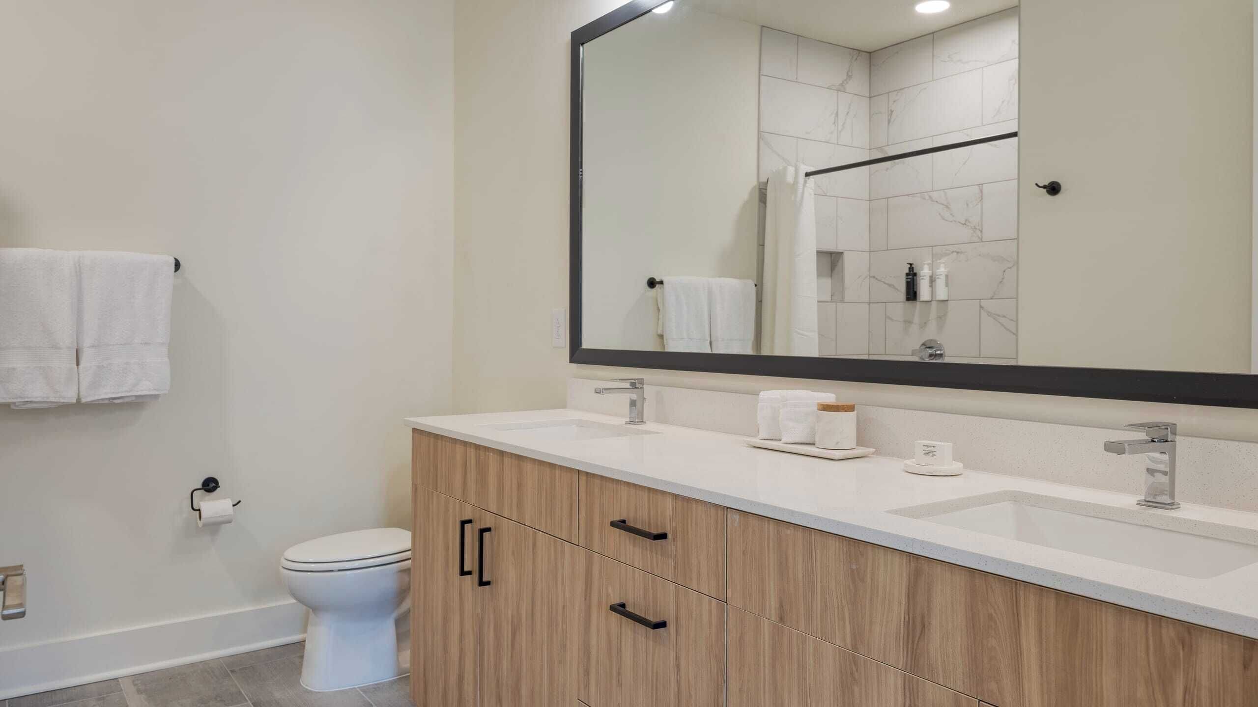 Ein modernes Badezimmer mit Toilette in Weiß, einem Holzbadezimmer-Möbelstück mit zwei Schubladen, einem großen Spiegel und einem weißen Waschbecken.