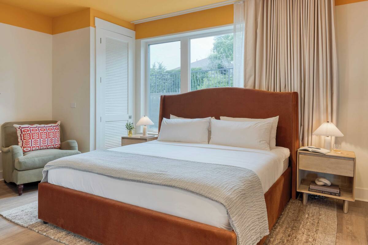 Une chambre à coucher avec un grand lit, une table de chevet avec une lampe, une chaise et une fenêtre avec des rideaux.