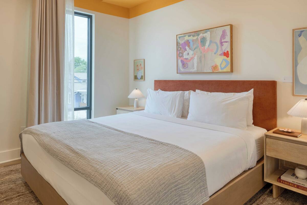 Une chambre d’hôtel avec un grand lit, une table de chevet avec une lampe, une bibliothèque et une fenêtre avec des rideaux.