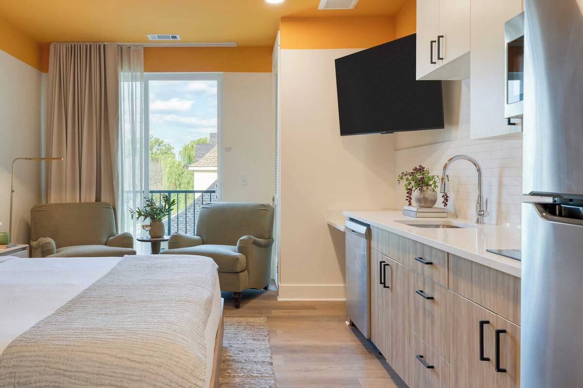 Une chambre d’hôtel comprenant un lit, une chaise, une petite table avec une plante, une fenêtre avec des rideaux et une kitchenette équipée de placards et d’un téléviseur.