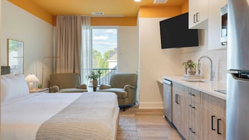Una habitación de hotel con una cama, una silla, una mesita con una planta, una ventana con cortinas y una kitchenette con armarios y un televisor.