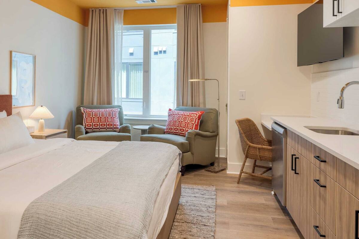 Une chambre d’hôtel avec un grand lit, deux fauteuils, une petite table, un canapé, une télévision, un coin cuisine avec un évier et des placards, et une fenêtre avec des rideaux.