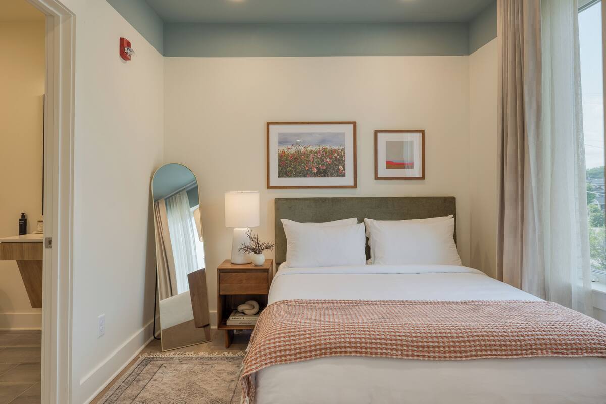 Une chambre d’hôtel bien rangée, avec un grand lit, une table de chevet avec une lampe, un tableau encadré et une fenêtre avec des rideaux.