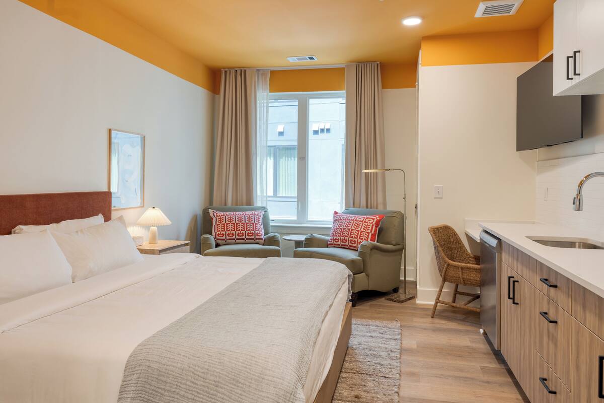 Une chambre d’hôtel comprenant un lit, deux fauteuils, un bureau, une chaise, une fenêtre et un coin cuisine.