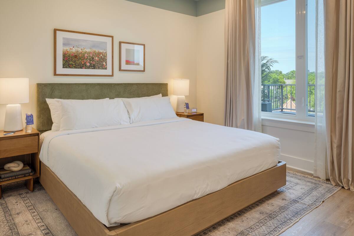 Une chambre d’hôtel avec un grand lit, deux tables de chevet, une petite table avec une lampe, un tableau encadré et une fenêtre avec des rideaux.