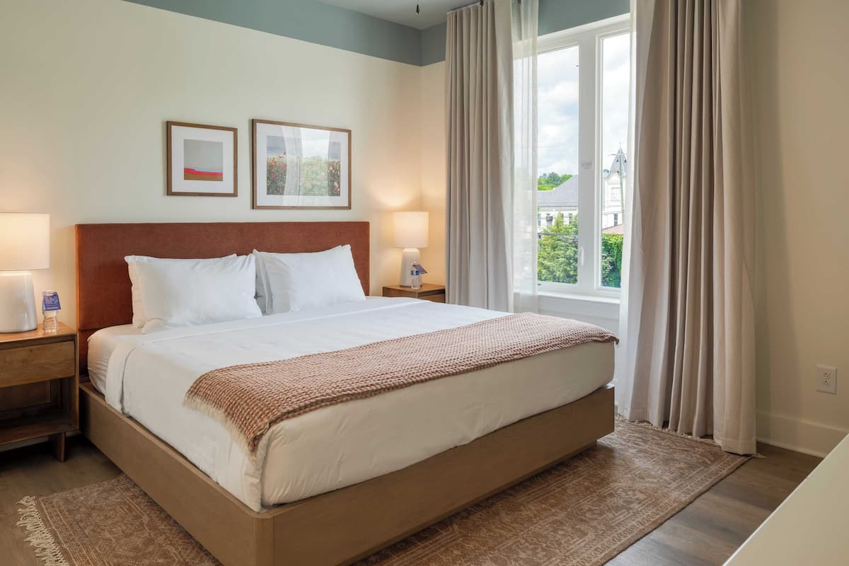 Une chambre d’hôtel avec un grand lit, deux tables de chevet, une petite table, une fenêtre avec des rideaux et deux tableaux encadrés accrochés au mur.