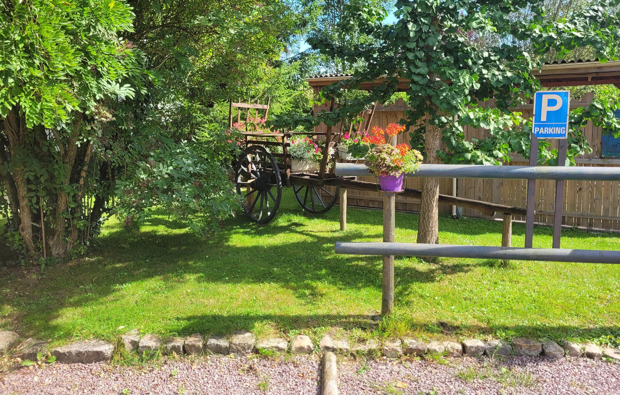 A wooden cart with flower pots parked under a tree, surrounded by greenery and a wooden fence.