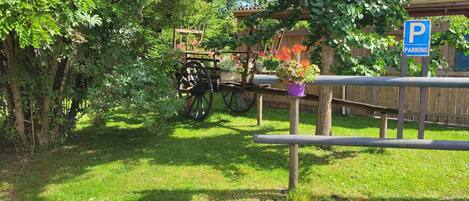 A wooden cart with flower pots parked under a tree, surrounded by greenery and a wooden fence.