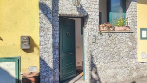 Una calle estrecha con un muro de piedra, una puerta verde, un buzón y una ventana con flores.