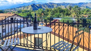Un balcón con una mesa, una botella de vino y una copa. El balcón ofrece vistas a un paisaje montañoso con vegetación y un pueblo al fondo.