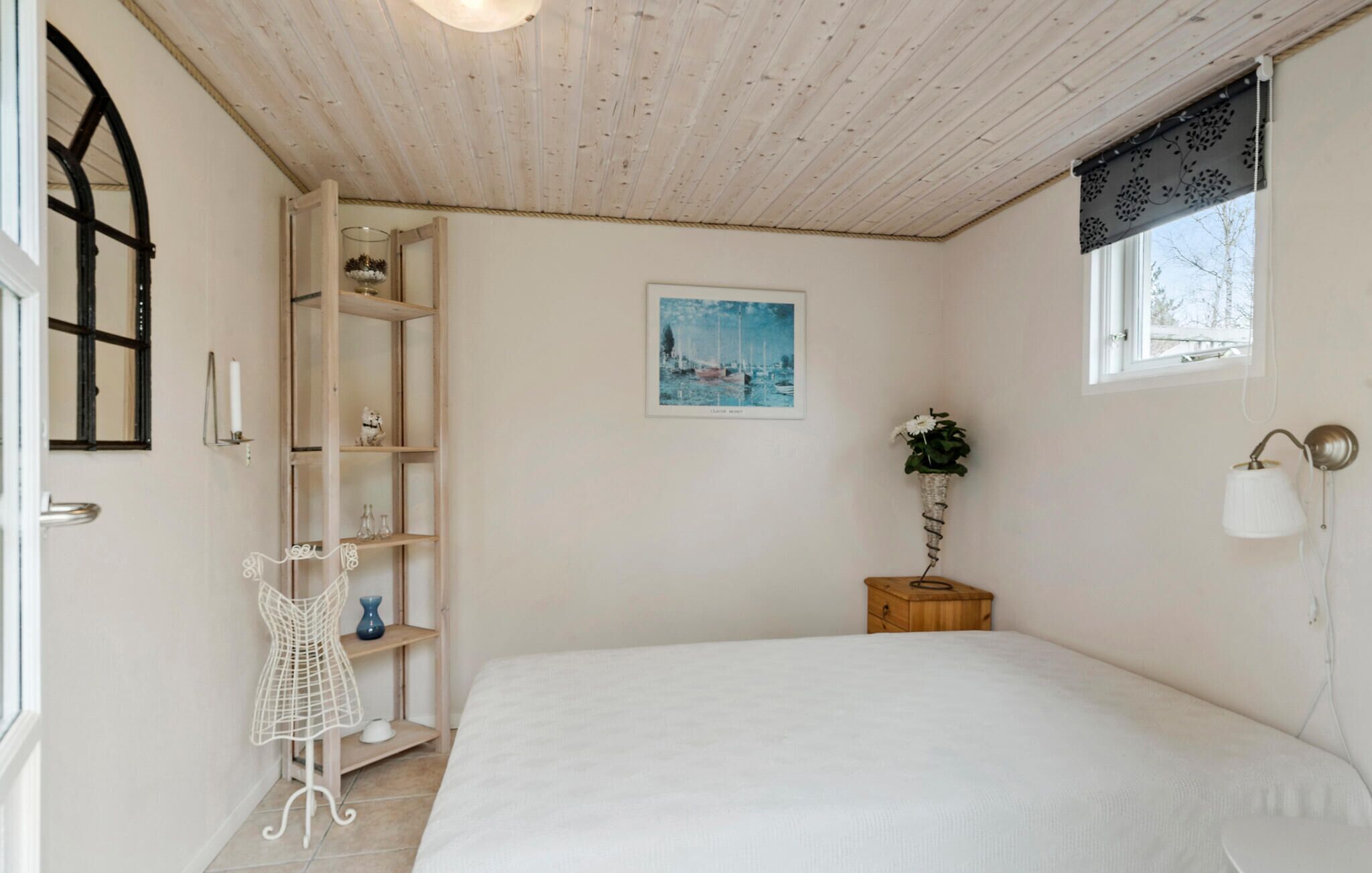 Une chambre avec un lit, une étagère en bois, une petite table avec une plante, une lampe fixée au mur et un tableau encadré.