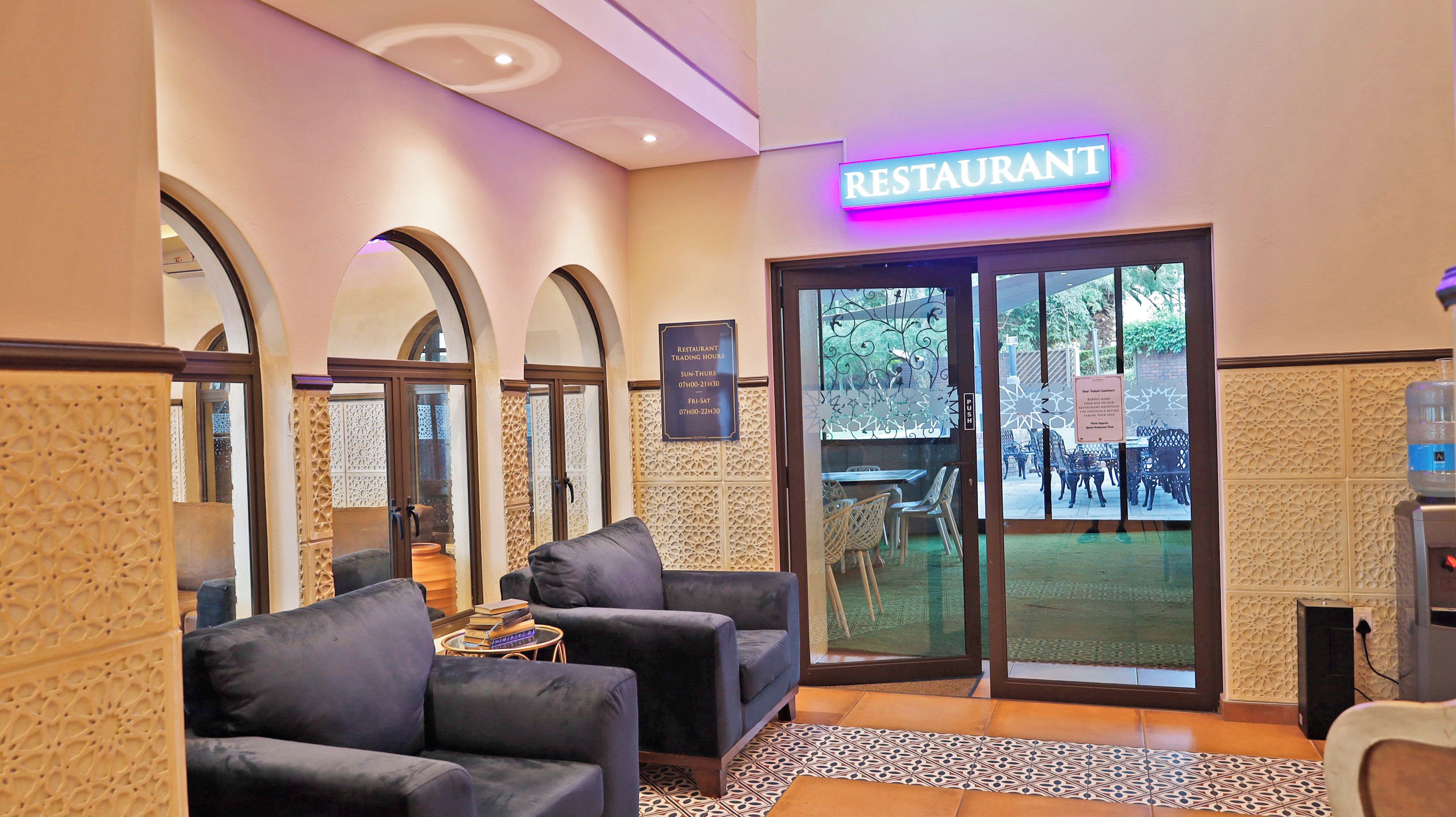 A restaurant with a neon sign, seating area with chairs, a water dispenser, and a patterned floor.