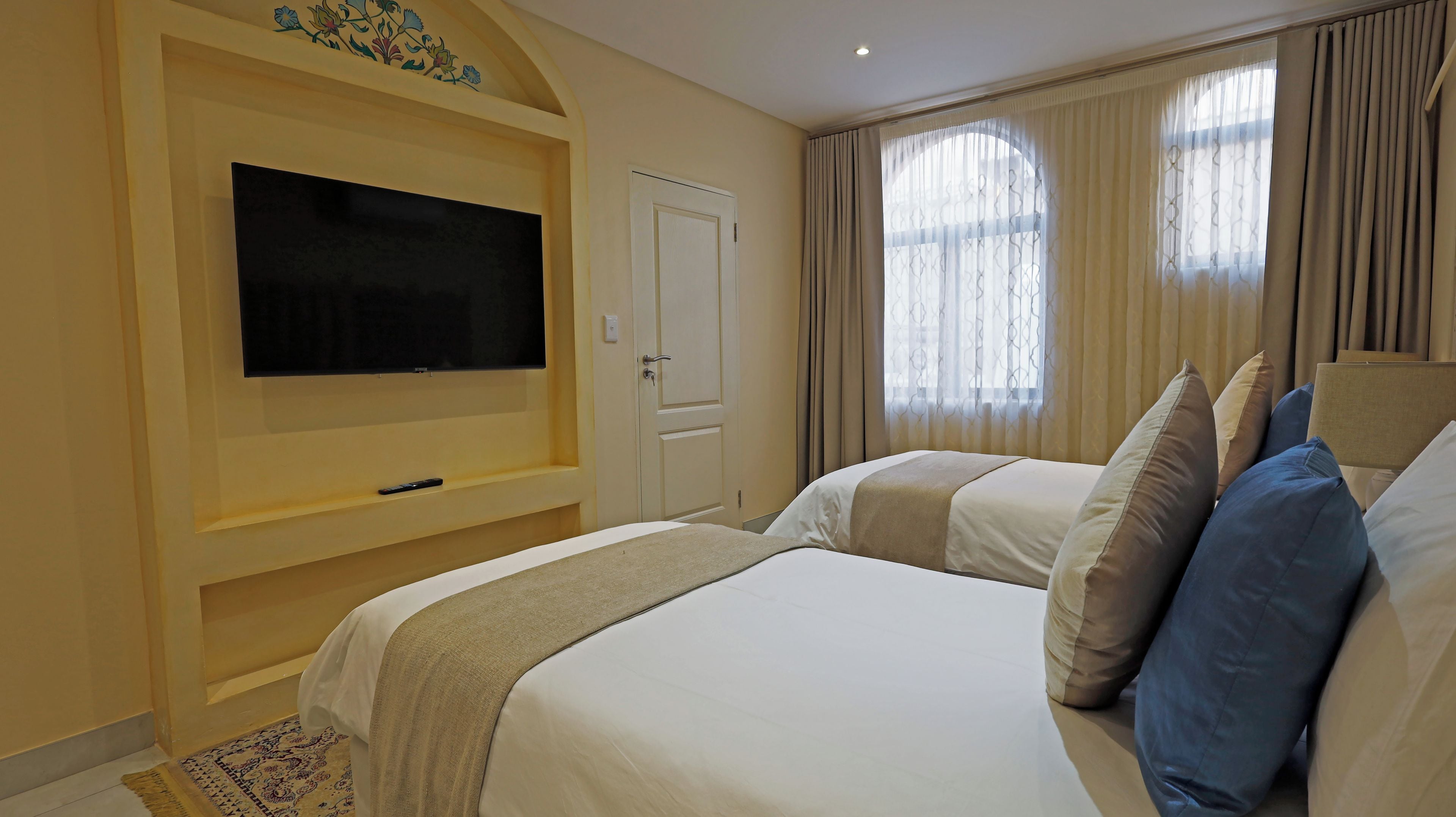 A hotel room with a large flat-screen TV mounted on the wall, a bed with multiple pillows, and a window with curtains.