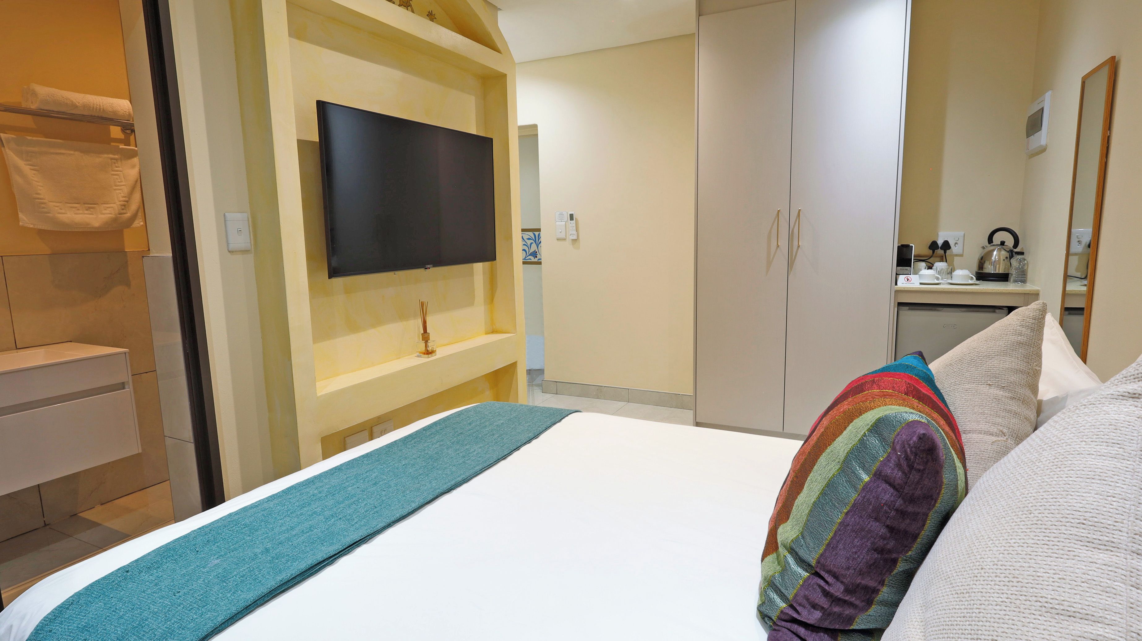 Habitación de hotel con cama, televisor montado en la pared, armario y una pequeña zona de kitchenette.