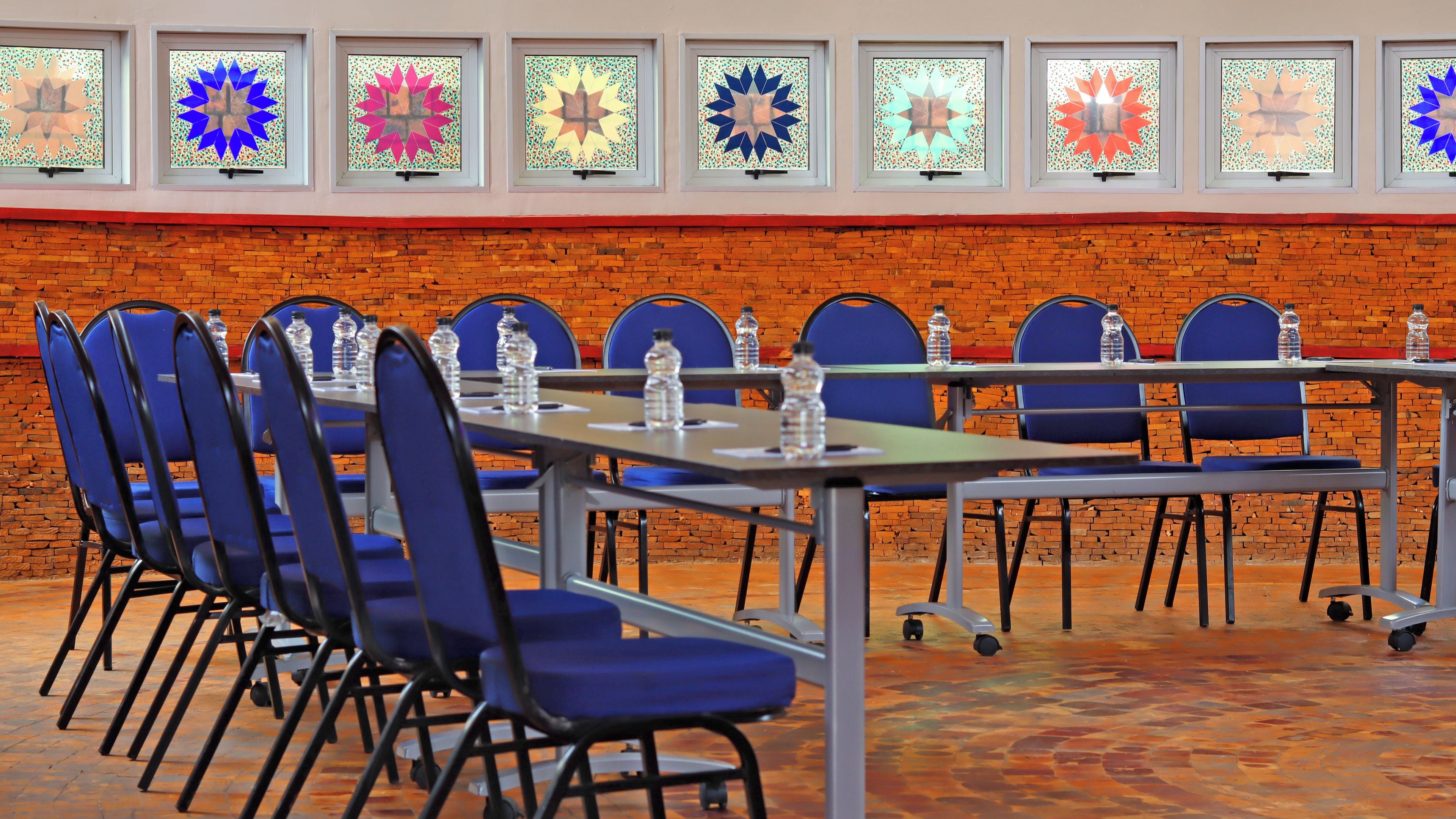 A conference room with a brick wall, blue chairs, tables with water bottles, and colorful stained glass windows.