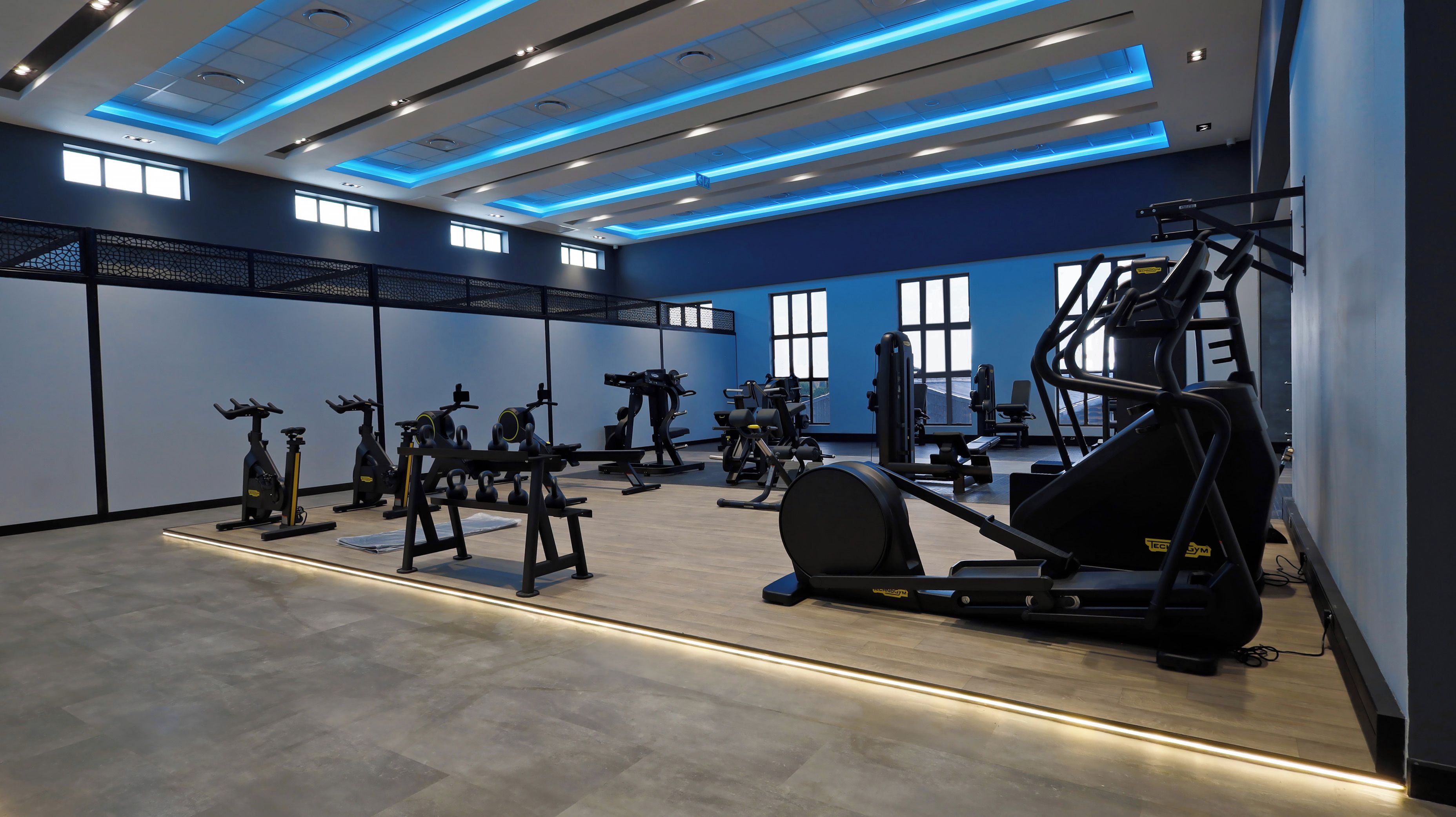 Un gimnasio moderno con equipos de ejercicio variados, ventanas amplias y techo iluminado en azul.