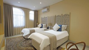 A hotel room with two beds, a patterned rug, a decorative headboard, and an air conditioning unit. - Qurtuba Resort And Events (Johannesburg)