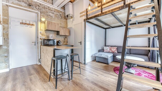 Un apartament modern d'estil loft amb parets de maó al descobert, una zona de cuina, un saló amb sofà i taula de centre, i un llit d'altitud accessible per una escala de fusta.