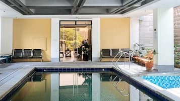 Una piscina interior con vista despejada al área de la piscina, una puerta que conduce a otra habitación, y algunas sillas y plantas en macetas.