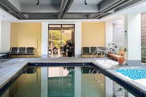 Una piscina interior con vista despejada al área de la piscina, una puerta que conduce a otra habitación, y algunas sillas y plantas en macetas.
