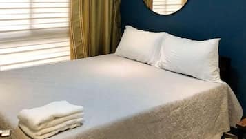 Una cama bien hecha con una toalla blanca doblada, un espejo redondo en la pared y una ventana con persianas.