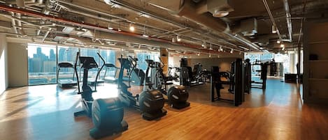A spacious gym with various exercise equipment, large windows offering city views, and exposed ductwork on the ceiling.